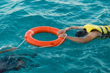 A bearded man diver in a life jacket holds on to a lifebuoy in transparent azure sea water.