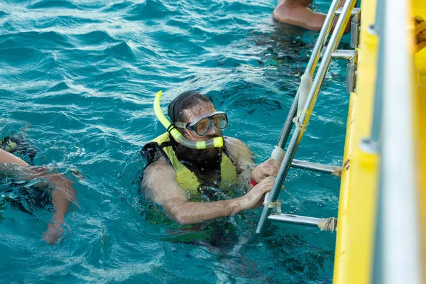 A bearded man diver in a life jacket climbs on board a boat in the sea.