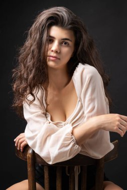Classic studio portrait of a young brunette in a white loose top, who is sitting on a chair against a black background.
