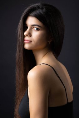 Classic studio portrait of a young brunette dressed in a black top, who is sitting on a chair against a black background.