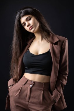 Classic studio portrait of a young brunette dressed in a black top and formal suit, who is sitting on a chair against a black background.
