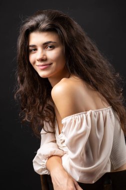Classic studio portrait of a young brunette in a white loose top, who is sitting on a chair against a black background.