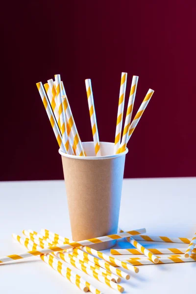 Disposable cardboard biodegradable cup on a white table with a straw against a red background.