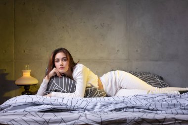 A young attractive girl dressed in white pajamas is resting on a bed in a bedroom in the evening lighting.