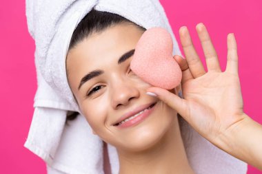 Beautiful cheerful attractive girl with a towel on her head, holds a sponge in the form of a pink heart.