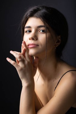 Classic studio portrait of a young brunette dressed in a black top, who is sitting on a chair against a black background.