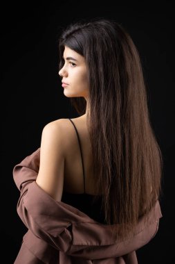 Classic studio portrait of a young brunette dressed in a black top and formal suit, who is sitting on a chair against a black background.