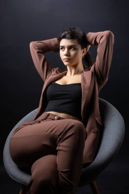 Klasik bir stüdyo portresi, siyah bir arka planda bir sandalyede oturan, siyah bluz ve resmi bir takım giymiş genç bir esmer..
