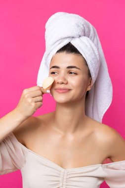 Beautiful cheerful attractive girl with a towel on her head, holds a sponge against a pink background.