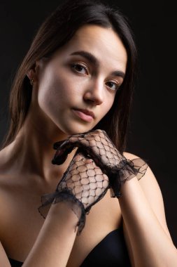 Classic dark studio portrait of a young brunette woman in black clothes who is sitting on a chair.