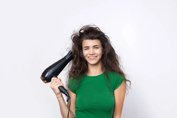 stock image A cute young girl dressed in a green top dries her beautiful long silky hair with a hair dryer against a white background.