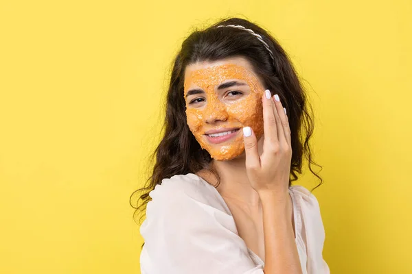 A young brunette girl with a cleansing scrub mask on her face, looks at the camera and smiles, against a yellow background.