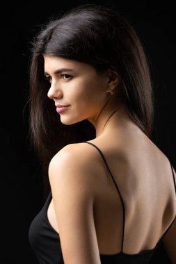 Classic studio portrait of a young brunette dressed in a black top, who is sitting on a chair against a black background.