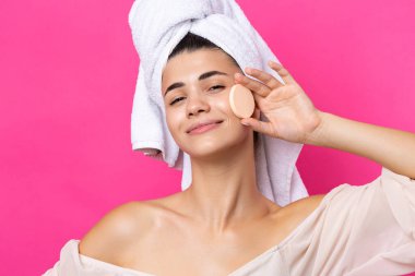 Beautiful cheerful attractive girl with a towel on her head, holds a sponge against a pink background.