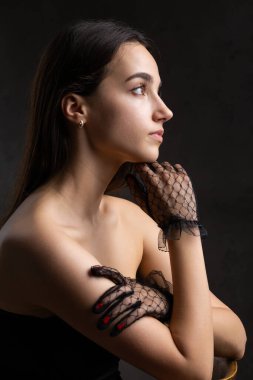 Classic dark studio portrait of a young brunette woman in black clothes who is sitting on a chair.