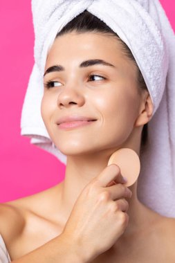 Beautiful cheerful attractive girl with a towel on her head, holds a sponge against a pink background.