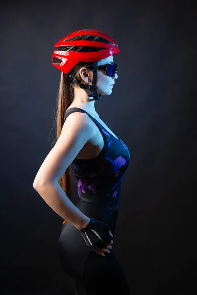 stock image A young female cyclist wearing a safety helmet and glasses, dressed in a bib shorts poses against a black background in the studio.