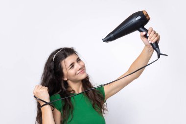 A cute young girl dressed in a green top dries her beautiful long silky hair with a hair dryer against a white background.