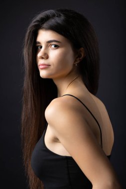 Classic studio portrait of a young brunette dressed in a black top, who is sitting on a chair against a black background.