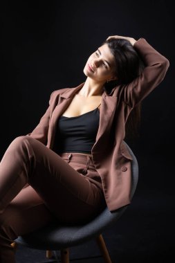 Classic studio portrait of a young brunette dressed in a black top and formal suit, who is sitting on a chair against a black background.