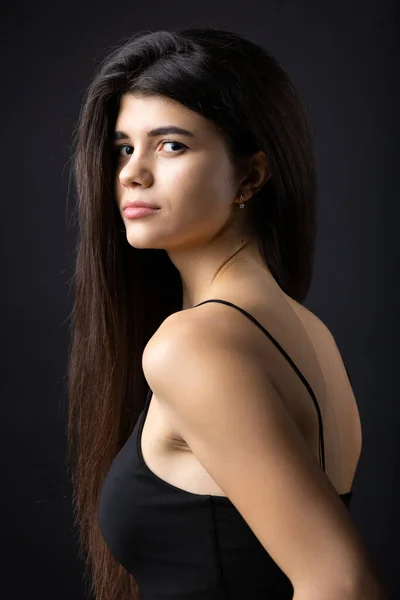 Classic studio portrait of a young brunette dressed in a black top, who is sitting on a chair against a black background.