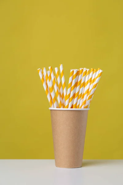 Disposable cardboard biodegradable cup on a white table with a straw against a yellow background.
