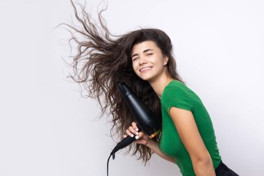 A cute young girl dressed in a green top dries her beautiful long silky hair with a hair dryer against a white background.