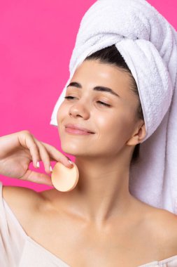 Beautiful cheerful attractive girl with a towel on her head, holds a sponge against a pink background.