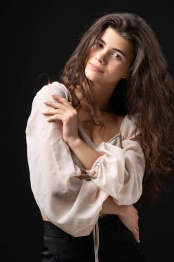Classic studio portrait of a young brunette in a white loose top, who is sitting on a chair against a black background.