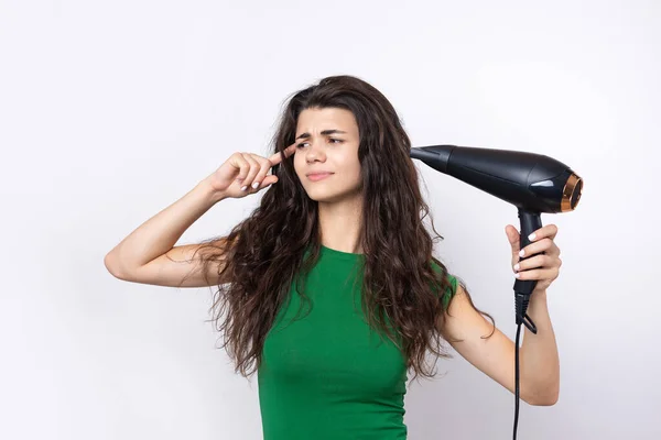 A cute young girl dressed in a green top dries her beautiful long silky hair with a hair dryer against a white background.