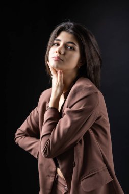 Classic studio portrait of a young brunette dressed in a black top and formal suit, who is sitting on a chair against a black background.
