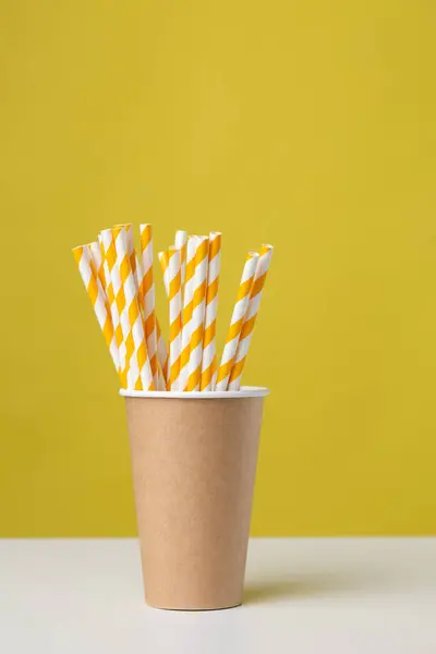 Disposable cardboard biodegradable cup on a white table with a straw against a yellow background.