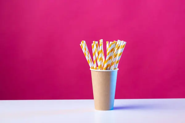 Disposable cardboard biodegradable cup on a white table with a straw against a red background.