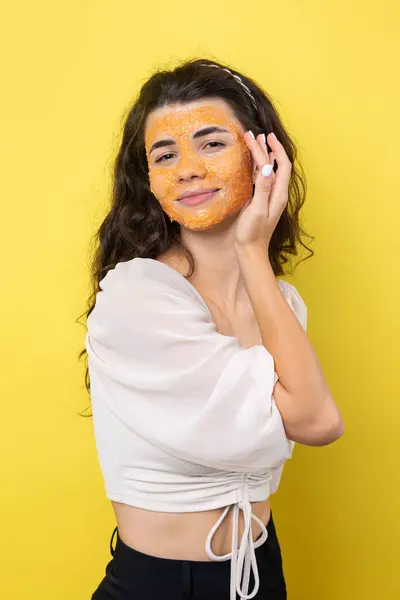 A young brunette girl with a cleansing scrub mask on her face, looks at the camera and smiles, against a yellow background.