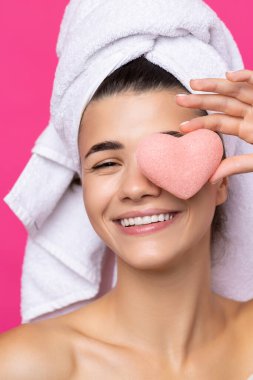 Beautiful cheerful attractive girl with a towel on her head, holds a sponge in the form of a pink heart.