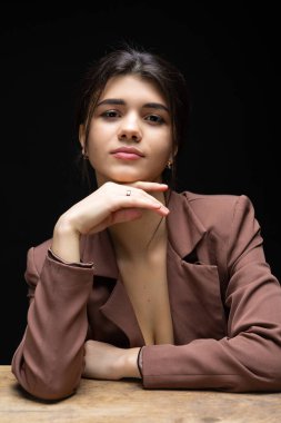 Classic studio portrait of a young brunette dressed in a black top and formal suit, who is sitting on a chair against a black background.