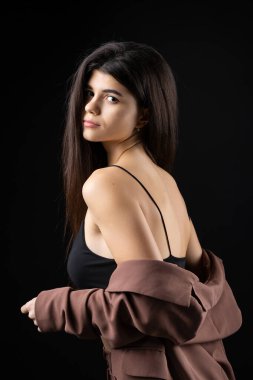 Classic studio portrait of a young brunette dressed in a black top and formal suit, who is sitting on a chair against a black background.