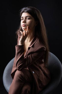 Classic studio portrait of a young brunette dressed in a black top and formal suit, who is sitting on a chair against a black background.