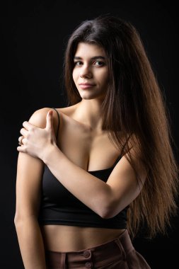 Classic studio portrait of a young brunette dressed in a black top, who is sitting on a chair against a black background.