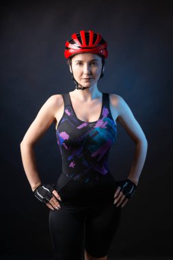 A young female cyclist wearing a safety helmet and glasses, dressed in a bib shorts poses against a black background in the studio.