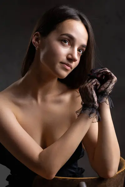 Classic dark studio portrait of a young brunette woman in black clothes who is sitting on a chair.