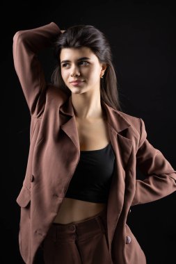 Classic studio portrait of a young brunette dressed in a black top and formal suit, who is sitting on a chair against a black background.