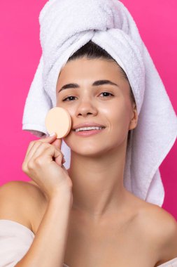 Beautiful cheerful attractive girl with a towel on her head, holds a sponge against a pink background.