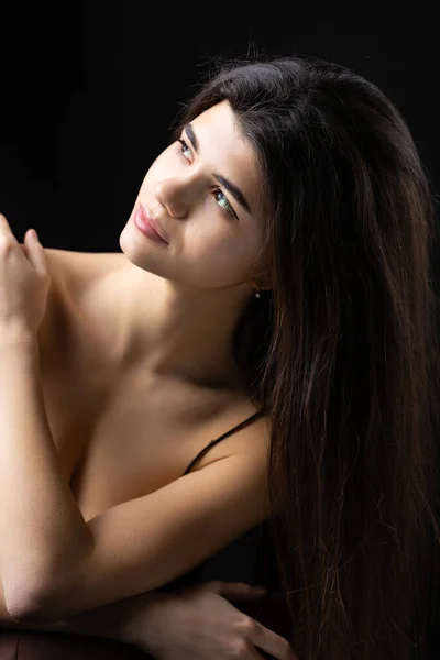 Classic studio portrait of a young brunette dressed in a black top, who is sitting on a chair against a black background.