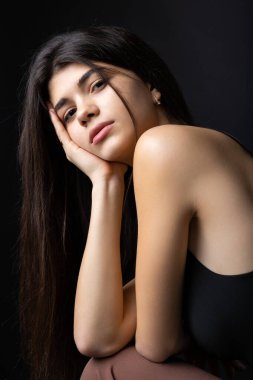 Classic studio portrait of a young brunette dressed in a black top, who is sitting on a chair against a black background.