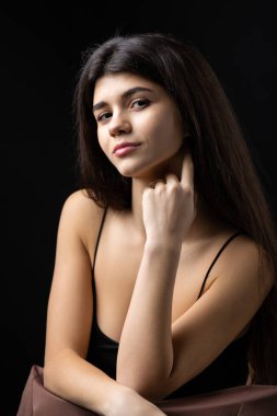Classic studio portrait of a young brunette dressed in a black top, who is sitting on a chair against a black background.