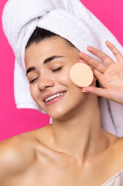 Beautiful cheerful attractive girl with a towel on her head, holds a sponge against a pink background.