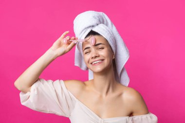 A beautiful cheerful attractive girl with a towel on her head holds a cosmetic roller near her face against a pink background.