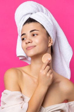 Beautiful cheerful attractive girl with a towel on her head, holds a sponge against a pink background.