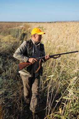 Mature man hunter with gun while walking on field.